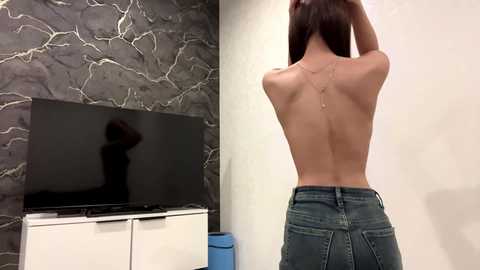 Video of a slender, topless woman with light skin, long brown hair, and a thin physique, wearing high-waisted jeans, standing in a modern, minimalist room with a dark marble wall and a flat-screen TV on a white cabinet.
