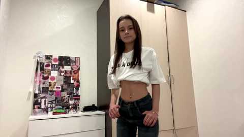 Video of a young East Asian woman with straight black hair, wearing a white crop top and low-rise jeans, standing in a cluttered bedroom with a collage on the wall.