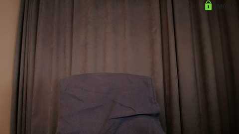 Media: A video of a dimly lit room with a beige curtain and a purple blanket on a chair in the foreground. The image has a green lock icon in the top right corner.