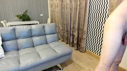 Video of a modern, light-gray, tufted leather sofa in a living room with beige walls, patterned curtains, and a wooden floor. A man's bare chest and arm are visible in the foreground.