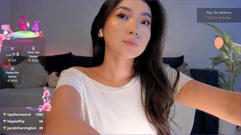 Video of a young East Asian woman with long black hair, wearing a white top, taking a selfie in a dimly lit living room with plants and a couch in the background.