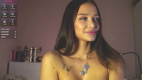 Video of a topless woman with long, straight, dark brown hair and light brown skin, wearing a silver necklace with a pendant. She has a serene expression. Background features a calendar, a bed, and a bookshelf.