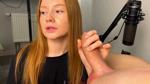 Video of a fair-skinned, red-haired young woman with a neutral expression, holding a microphone close to a circumcised, erect penis on a black stand. Background shows a white radiator and a white wall.