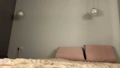 Video of a minimalist bedroom with a beige wall, two pink pillows, and two hanging spherical lights. The bed has a light-colored, patterned comforter.