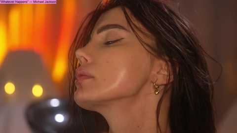 Video of a woman with long, wet, dark hair and closed eyes, wearing a gold hoop earring, set against a blurred, warm-toned cityscape background.