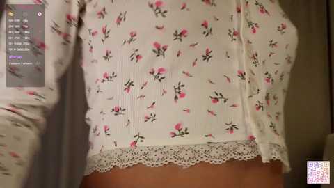 Video of a woman wearing a white, ribbed, long-sleeve top with pink floral patterns and lace trim. Background shows a dark room with a smartphone screen displaying a social media app.