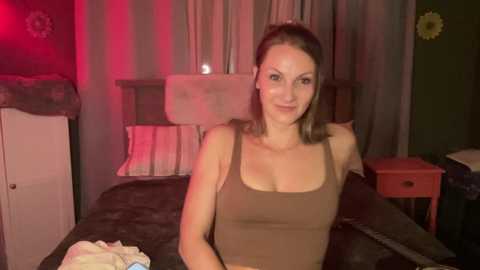 Video of a smiling Caucasian woman with light brown hair, wearing a brown tank top, sitting on a bed with a gray blanket, dimly lit room with red light, nightstand, and floral decorations.