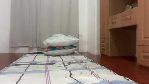 Video of a small, minimalist bedroom with a wooden bunk bed, a plaid blanket, and a stack of pillows against a wall.