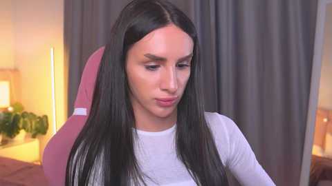 Video of a woman with long, straight black hair, wearing a white top, sitting on a pink gaming chair in a softly lit room with gray curtains.