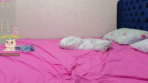 A video of a messy bedroom with a pink bedspread, two pillows, and a discarded pair of jeans on the floor. The room has a blue, tufted headboard and a pale pink wall.