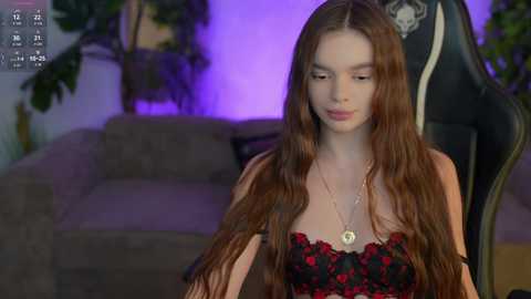 Video of a young woman with long, wavy brown hair, wearing a red and black floral strapless dress, sitting in a gaming chair with plants and a sofa in the background.