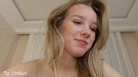 Video of a young, fair-skinned woman with blonde hair and a confident smile, wearing a pearl necklace, in a neutral-toned room with white curtains.