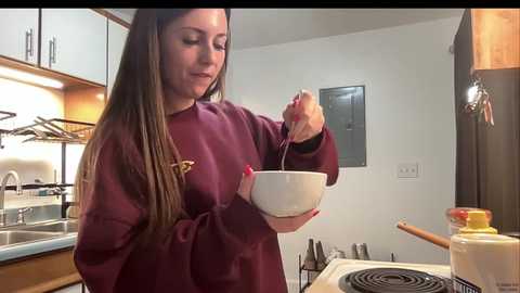 Video of a smiling young woman with long brown hair, wearing a maroon sweatshirt, stirring a white bowl with a spoon in a modern kitchen with stainless steel appliances and wooden cabinets.