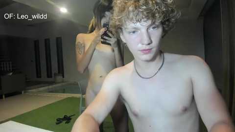 A video shows a young, shirtless, fair-skinned boy with curly hair, taking a selfie in a dimly-lit room, with a partially visible tattoo on his left shoulder.