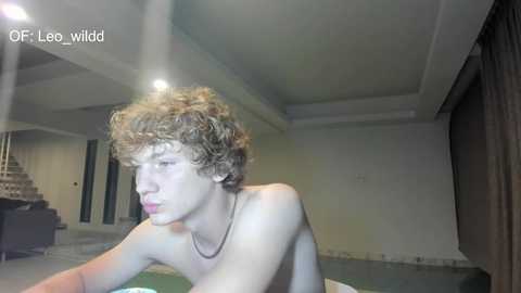 A video of a shirtless young man with curly, light brown hair, resting his head on a table in a dimly lit room with beige walls and a staircase in the background.
