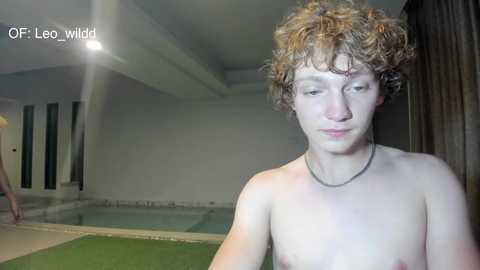 Video of a shirtless, curly-haired young man with fair skin, wearing a necklace, standing near a swimming pool in a dimly lit indoor setting.