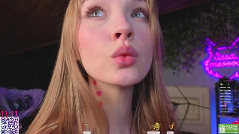 Video of a fair-skinned, blonde woman with blue eyes, puckered lips, and red dots on her neck, against a dimly lit room with neon signs and a video game console.