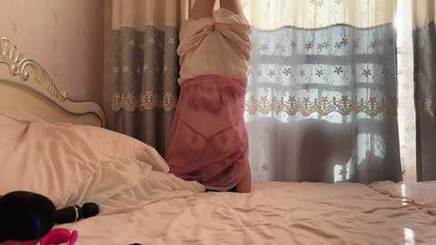 Video of a woman in a sheer pink dress, standing in a bedroom with white curtains and a bed.
