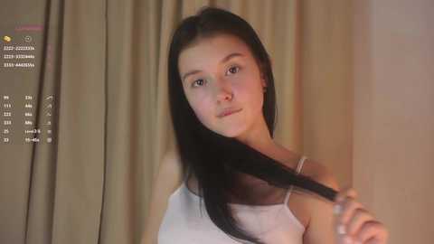 A video of a young woman with long black hair, fair skin, and a slim build, holding her hair up. She wears a white tank top. Background includes beige curtains and a digital calendar.