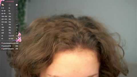 A video of a person with voluminous, wavy, shoulder-length brown hair, seen from the top down. The background is blurred, showing a green plant.