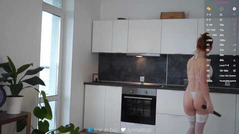 Video of a fair-skinned woman with red hair wearing white lingerie, standing in a modern kitchen with white cabinets, dark countertops, and a stove.