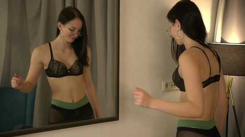 Video of a slender, fair-skinned woman with long dark hair, wearing black lace bra and green panties, smiling at her reflection in a mirror. Room features a lamp and gray curtains.