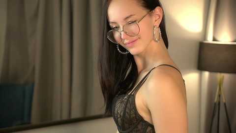 Video of a slender, fair-skinned woman with straight, dark hair, wearing round glasses and a black lace top, smiling in a softly lit room with beige curtains and a lamp.
