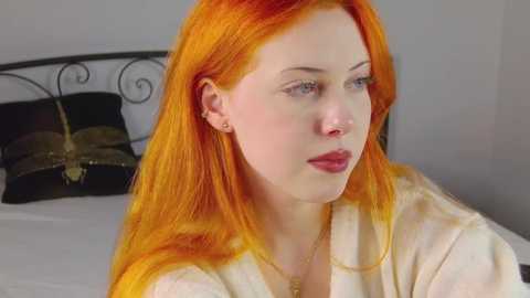 Media: Video of a fair-skinned woman with striking orange hair, wearing a white robe, sitting on a bed with a black headboard, in a simple bedroom.