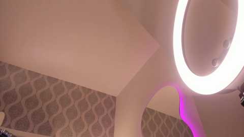 A video of a modern room with a large, circular, glowing white light fixture on the ceiling, casting a soft purple hue on the textured, patterned wallpaper.