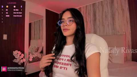 Video of a young woman with long black hair, wearing glasses and a white T-shirt with a motivational quote, seated on a white chair in a modern bedroom with white curtains and floral decor.