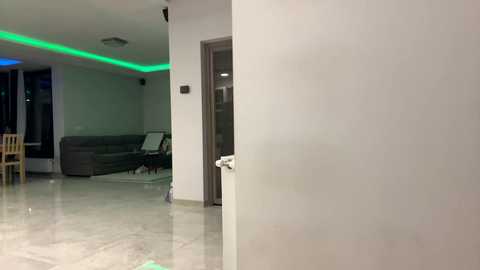 A video of a modern, minimalist living room with white walls, glossy marble flooring, and a grey sectional sofa. Green LED lights illuminate the ceiling, and a wooden chair is visible in the background.
