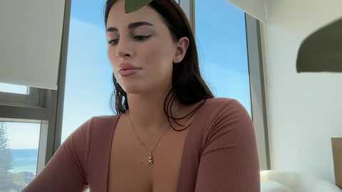 Video of a young woman with medium skin tone, dark hair, and a medium bust, wearing a pink ribbed top, standing indoors near large windows with a green leaf on her face.