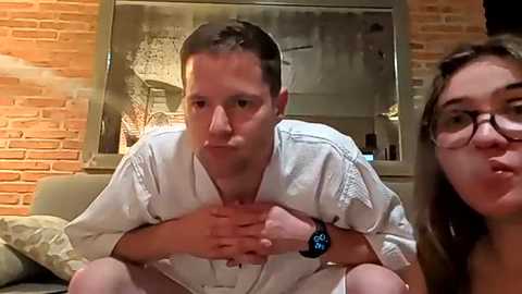 Video of two people in casual clothes, man with short hair and woman with glasses, sitting on a couch in a cozy living room with brick walls and a chandelier.