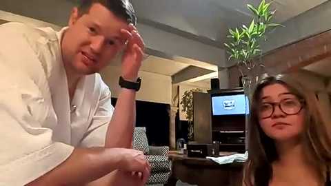 Video of a man with short hair in a white shirt, wearing a smartwatch, and a woman with long brown hair, wearing glasses, sitting on a couch in a modern living room.