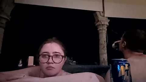 A video of a woman with glasses, lying on a bed, holding a can. The background shows a dark room with a column and a faint cityscape.