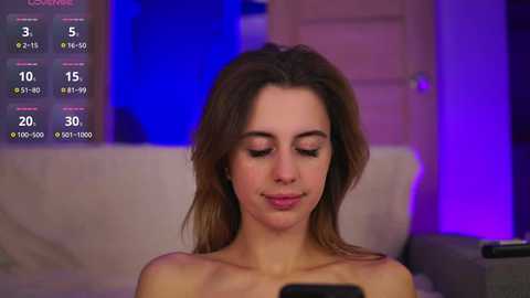 Media: Video of a young woman with light skin and long brown hair, wearing minimal makeup, sitting topless, looking at a smartphone, set in a dimly lit room with a white couch and purple lighting.