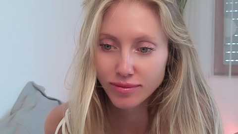 Video of a young woman with long, blonde hair, fair skin, and light makeup. She wears a sleeveless top. Background shows a gray pillow and a window with blinds.