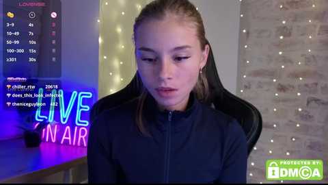Video of a young woman with light skin and long brown hair, wearing a black zip-up hoodie, sitting in a gaming chair with a brick wall and string lights background. Text overlays read \"Live In Air\" and \"Streamer by DMCA.\
