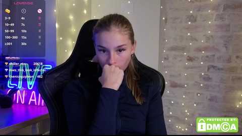Media: A video of a young woman with light skin and brown hair, wearing a black shirt, sitting with her hand on her mouth, in a dimly lit room with brick walls and fairy lights.