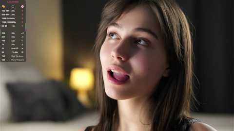 Video of a young Caucasian woman with straight, shoulder-length brown hair, looking up with parted lips and wide eyes, in a dimly lit bedroom with a blurred background.