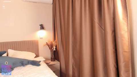 A video of a modern, minimalist bedroom with beige walls, a beige headboard, and a large, draped, taupe-colored curtain. The bed has white sheets and a blue pillow, with a small table beside it holding a vase of dried flowers.