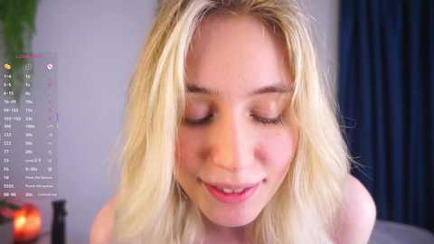 Media: A close-up video of a young, smiling, fair-skinned woman with long, blonde hair, wearing a white top, in a room with a calendar and dark curtains in the background.