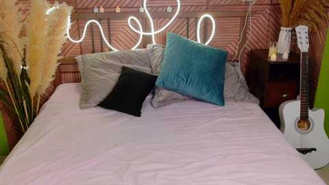 A cozy bedroom photo featuring a bed with a pink quilt, two pillows (one teal, one black), a guitar, a plant, and a glowing neon sign on a wooden headboard.