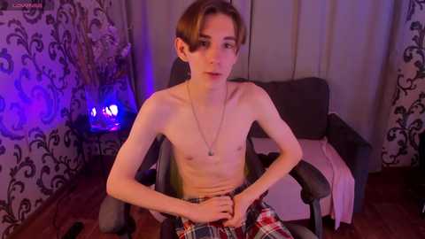 Video of a slender, shirtless teenage boy with light skin and short brown hair, seated in a chair, wearing plaid pants. Background features dark floral curtains, a purple-lit lamp, and a gray couch.