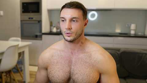 Video of a shirtless, muscular white man with short dark hair, light stubble, and a slight beard, sitting indoors in a modern kitchen with white cabinets, stainless steel appliances, and a dark gray couch.