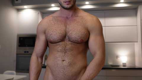 Video of a muscular, light-skinned, bearded man with a hairy chest and a small pubic area, standing naked in a modern kitchen with white cabinets and stainless steel appliances.