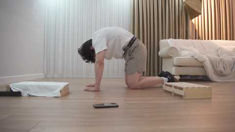 Video of a person with short dark hair, wearing a white T-shirt and beige shorts, kneeling on a light wooden floor, assembling furniture in a modern, light-colored room.