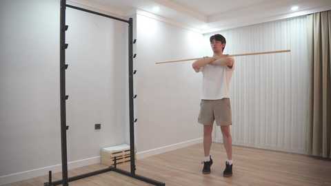 Video of a man in a white T-shirt and beige shorts doing a plank exercise on a wooden floor, with a black pull-up bar and a white weight bench in the background.