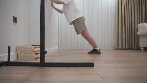 Video of a person in a white t-shirt and beige shorts standing on a wooden floor, adjusting a black tripod in a minimalist living room with white walls, beige curtains, and a cream-colored armchair.