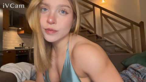 Video of a blonde, fair-skinned young woman with blue eyes, wearing a teal satin bra, seated on a couch in a cozy, dimly-lit living room with wooden staircase and kitchen background.
